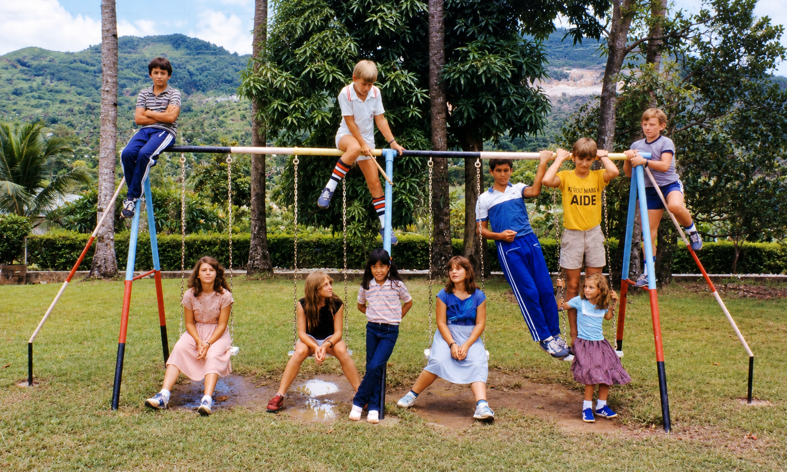 Schoolchildren sitting and standing on a swing set with green hills behind them.