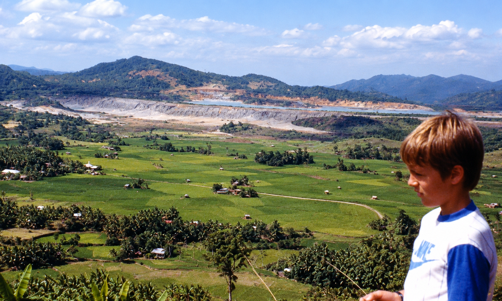 A boy overlooking green rice fields and the tailings pond from a hillside.