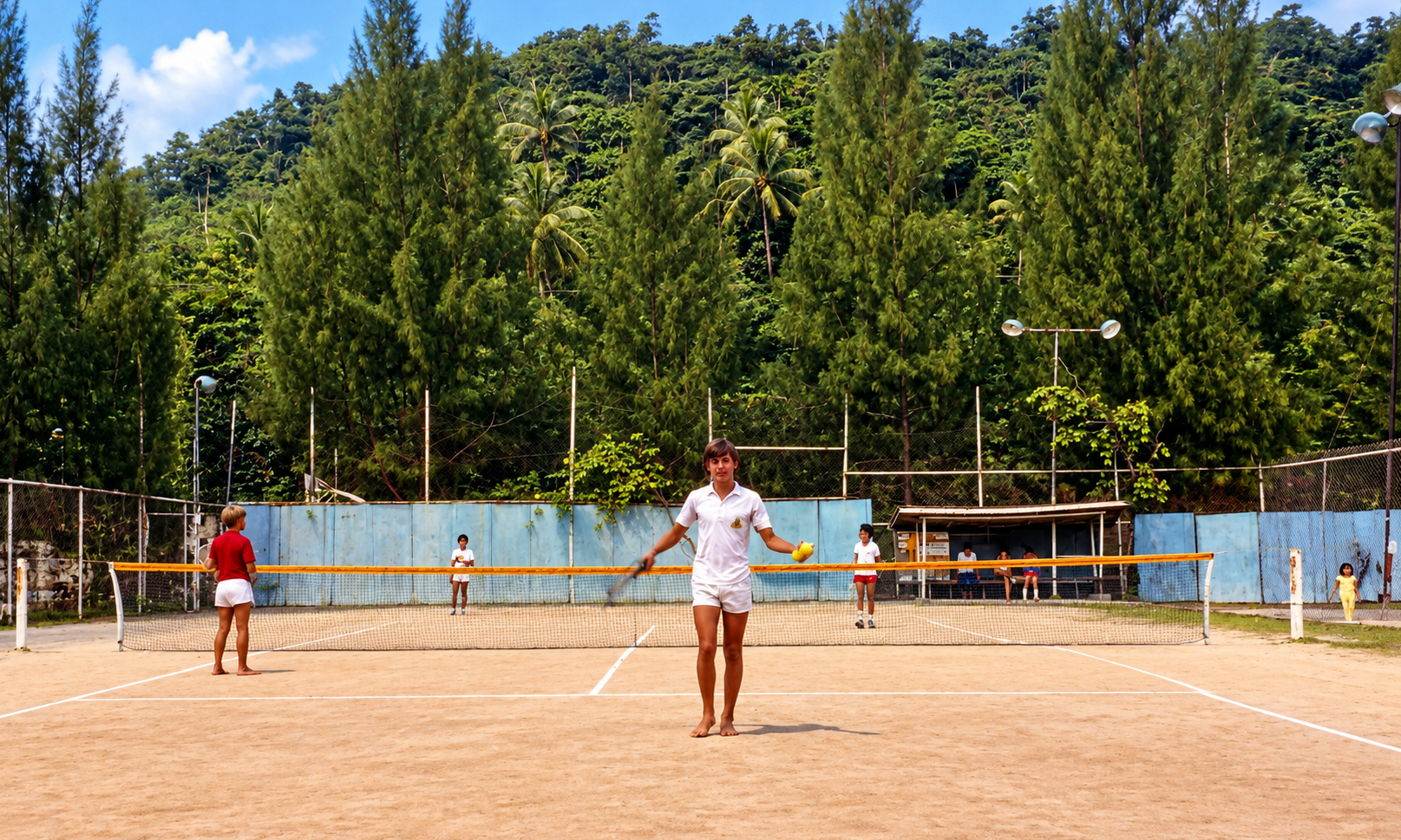 Players on a tennis court surrounded by tall trees and tropical vegetation.