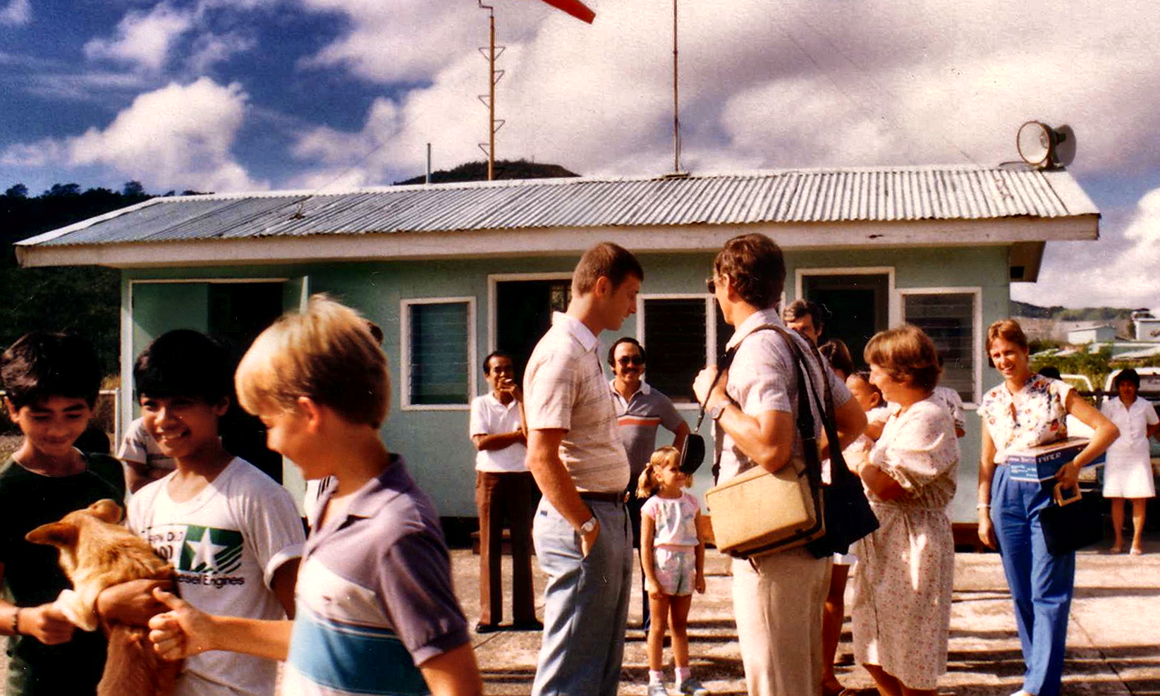 People gathered outside the airstrip terminal, with children and a dog in the foreground.