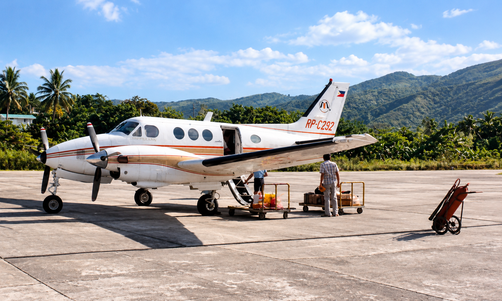 A white twin-engine plane with red and gold stripes beside baggage carts at the airstrip.