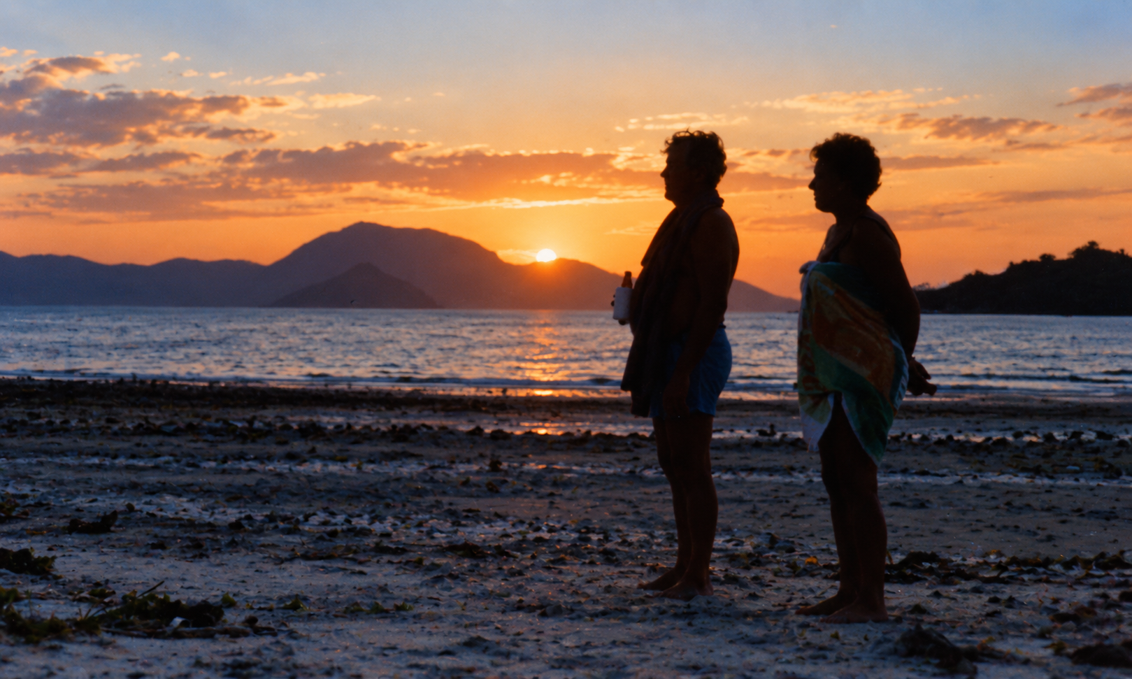 Two people standing on the beach as the sun sets behind the islands.