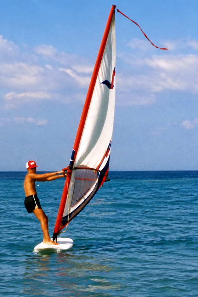 A windsurfer in a red cap balancing on a board with a tall white sail.