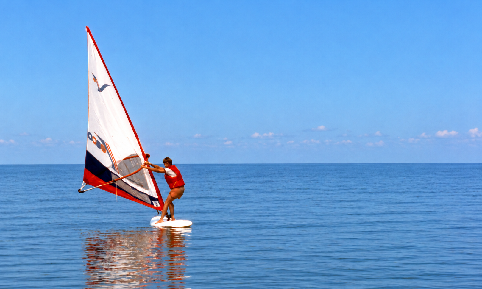 A windsurfer in a red life jacket crossing calm blue water.
