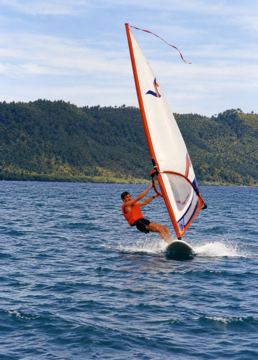 A windsurfer in an orange life jacket leaning back as the board cuts through the water.