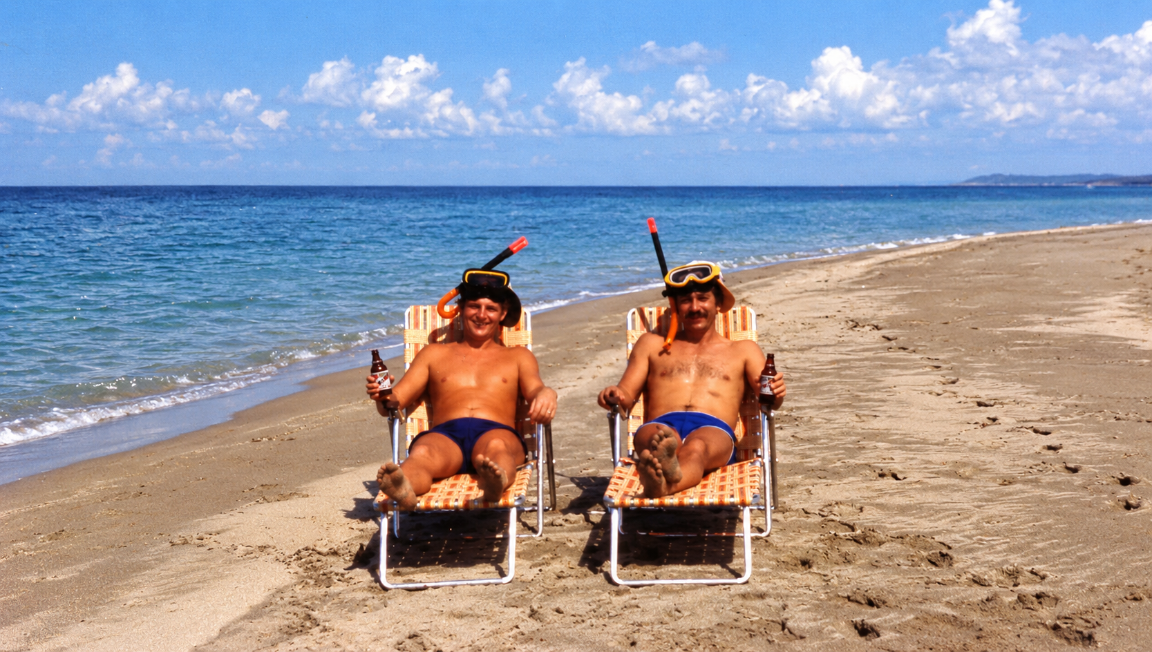 Two men wearing snorkeling masks and holding drinks while relaxing in beach chairs.