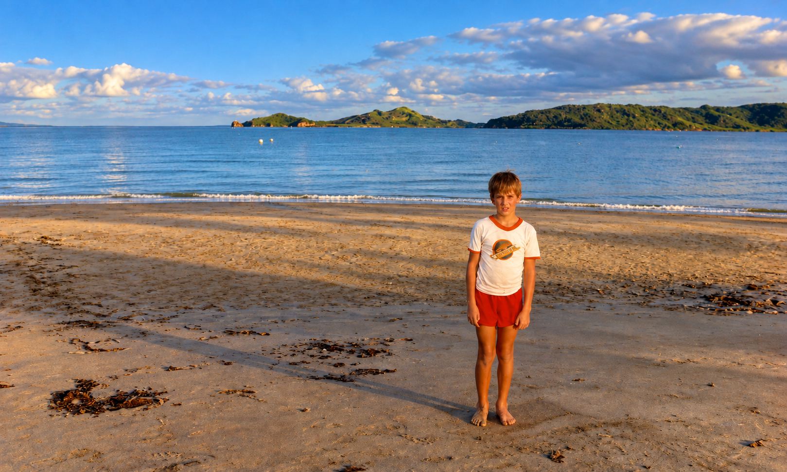A boy in a white shirt and red shorts standing on the beach in the evening light.