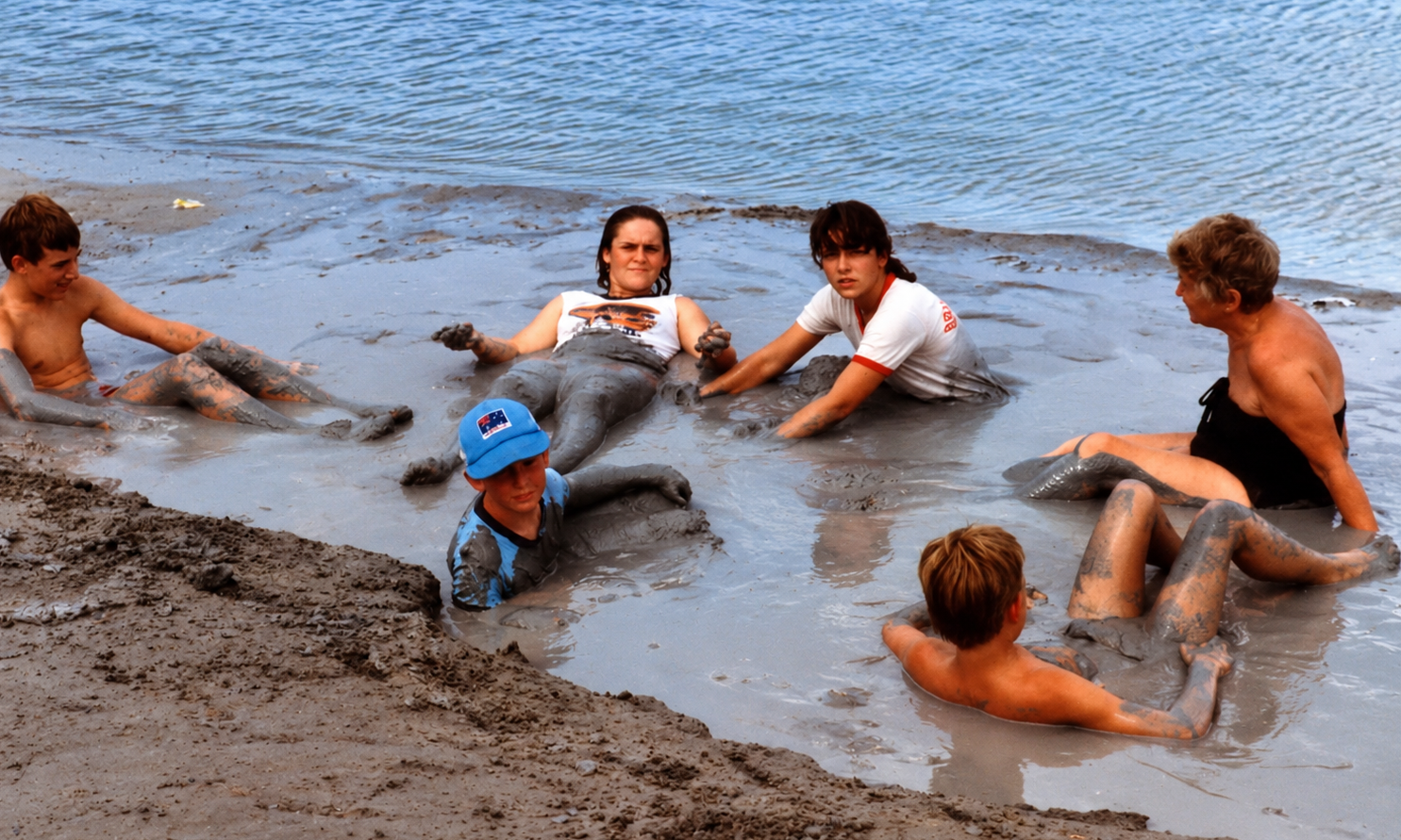 Adults and children sitting together in the soft mud beside the shoreline.