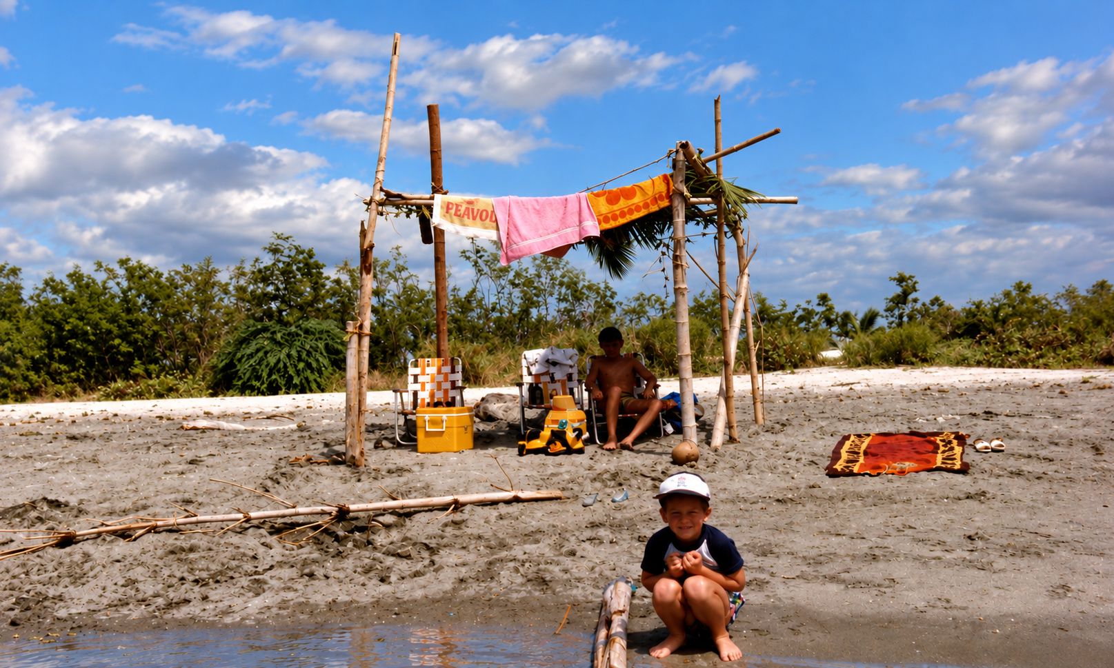 A child crouching near the water in front of a beach shelter hung with colorful towels.