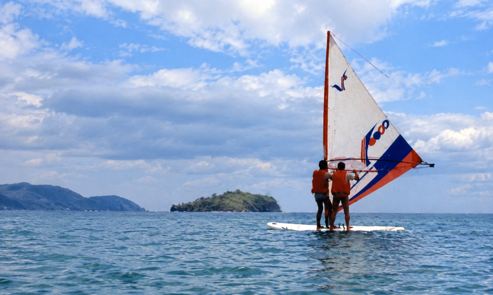 Two people in orange life jackets sharing a windsurfing board offshore.