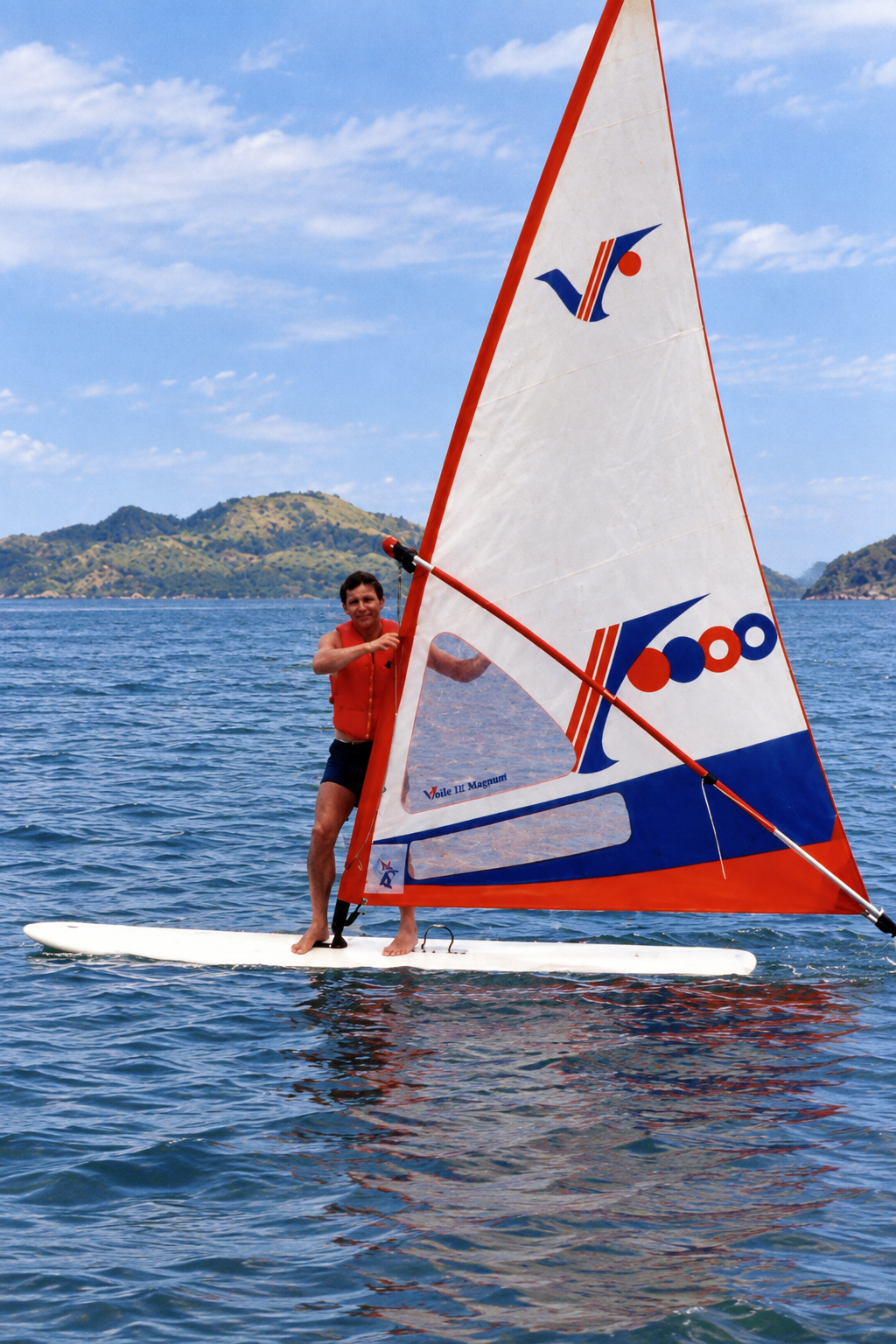A man in an orange life jacket standing on a windsurfing board with a red, white, and blue sail.
