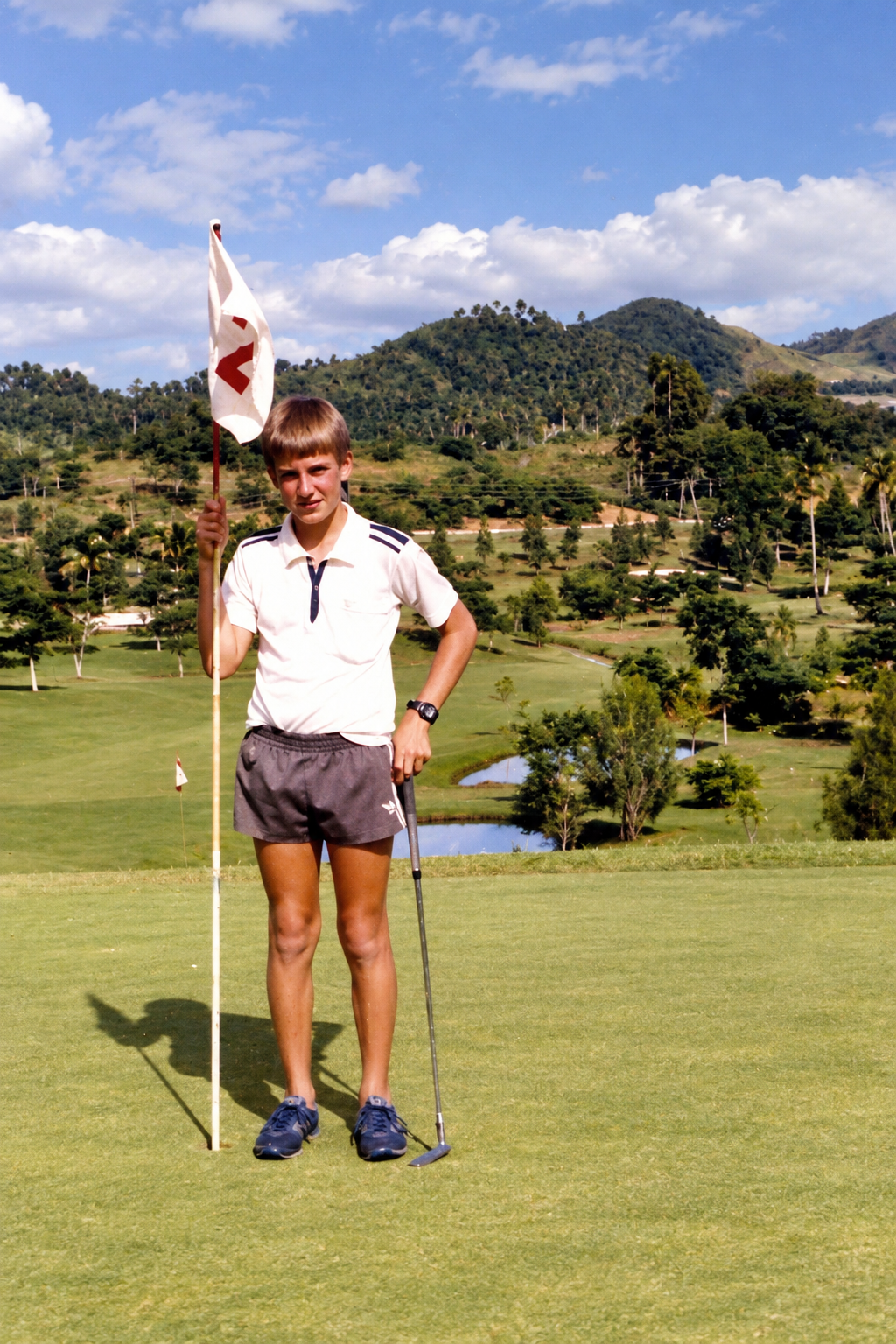 A young golfer holding a flagstick on the green, with hills and fairways behind him.