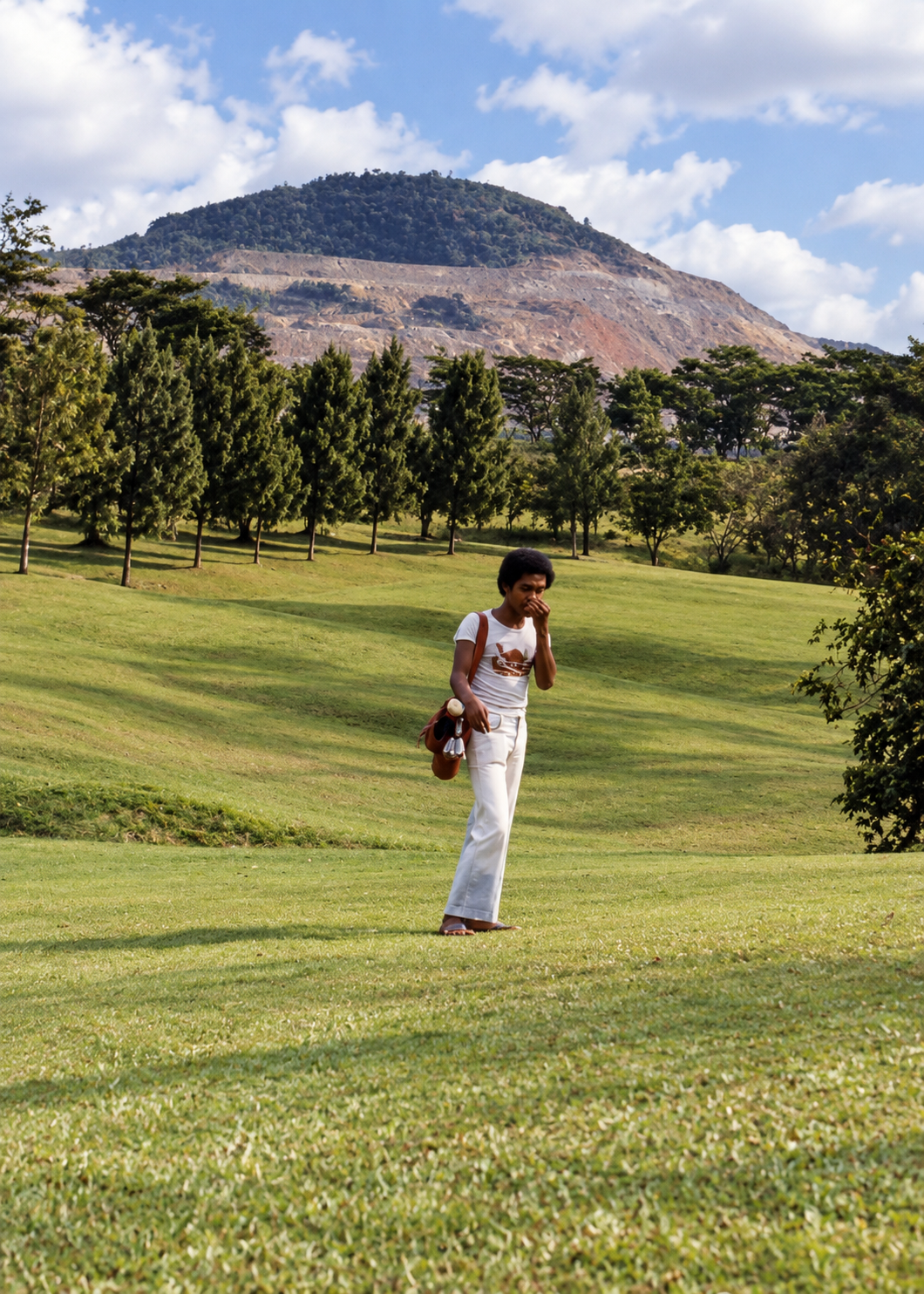 A caddie carrying golf clubs across the course, with the mine in the background.