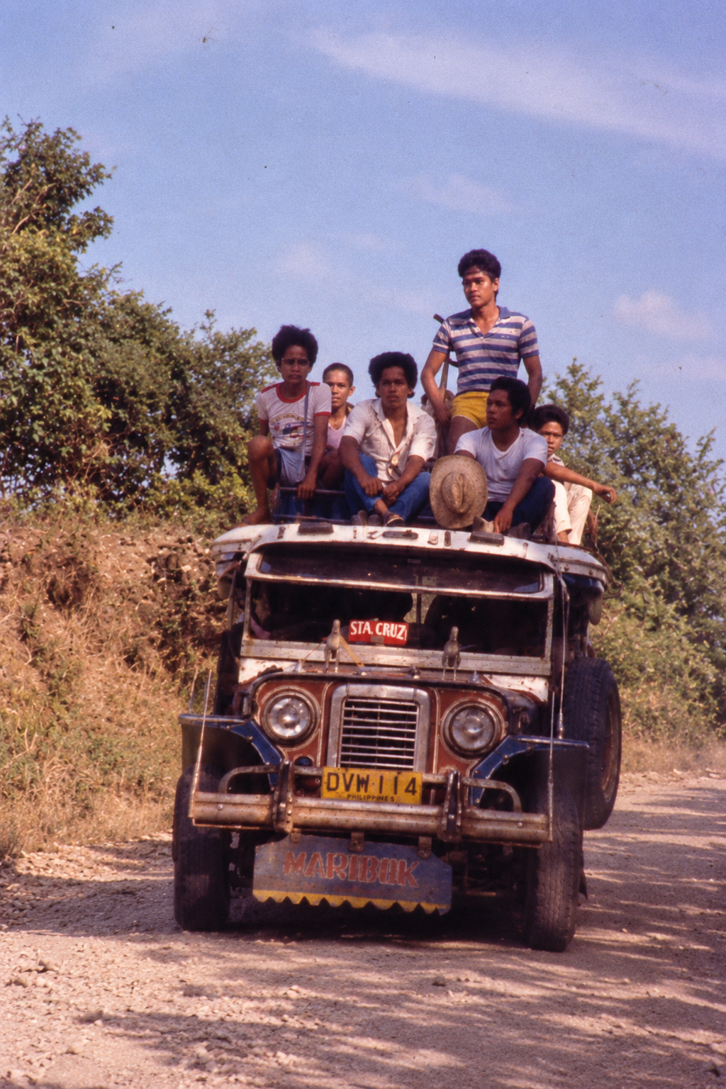 Passengers riding on the roof of a jeepney along a wooded dirt road.