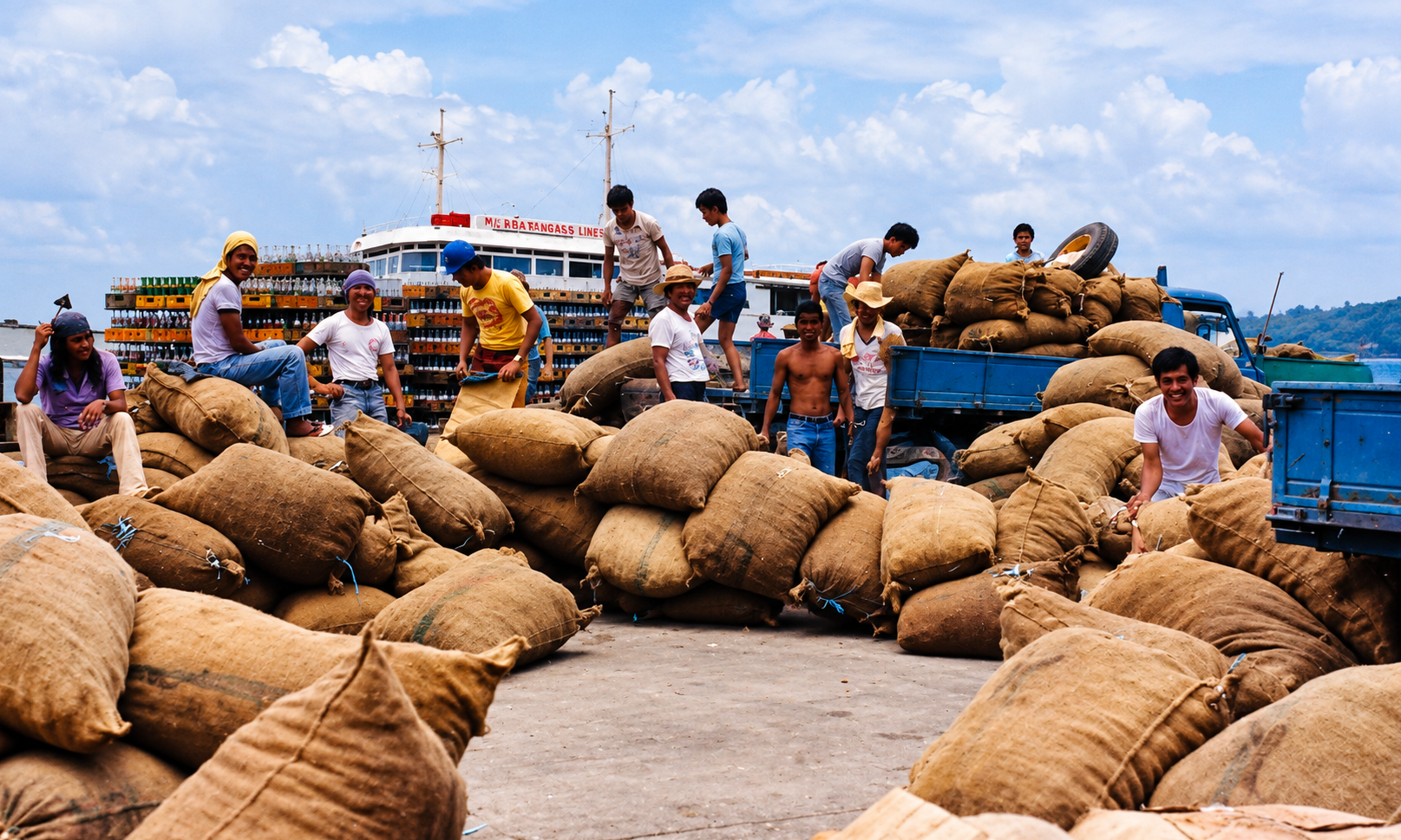 Workers among sacks and blue cargo trucks at a port, with a ferry behind them.