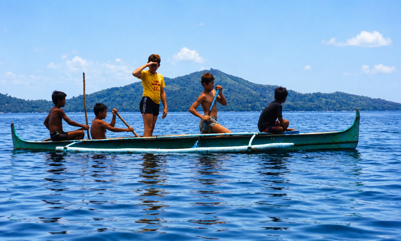 Five boys in an outrigger canoe on blue water with green hills in the distance.