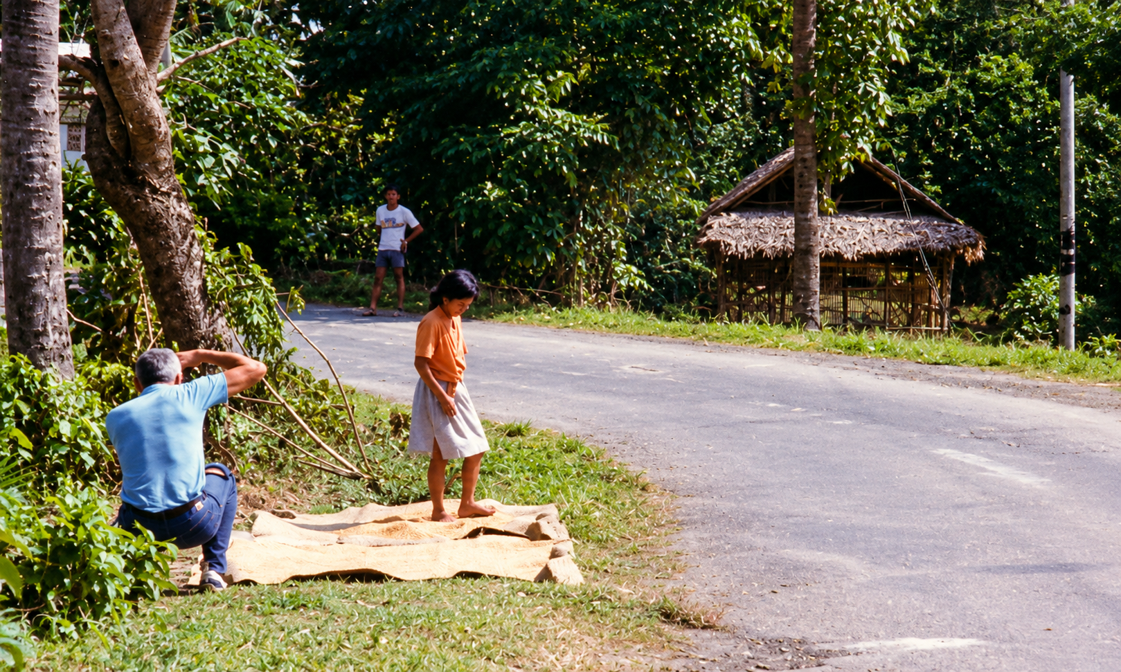 A man photographing a woman standing on grain drying beside a road.