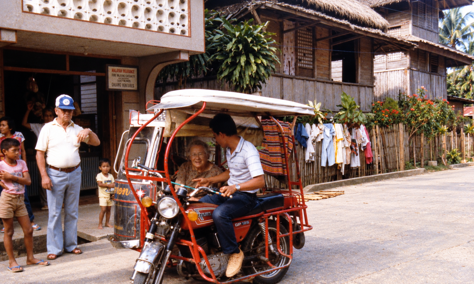 A woman riding in a red motorized tricycle beside a man standing on the street.