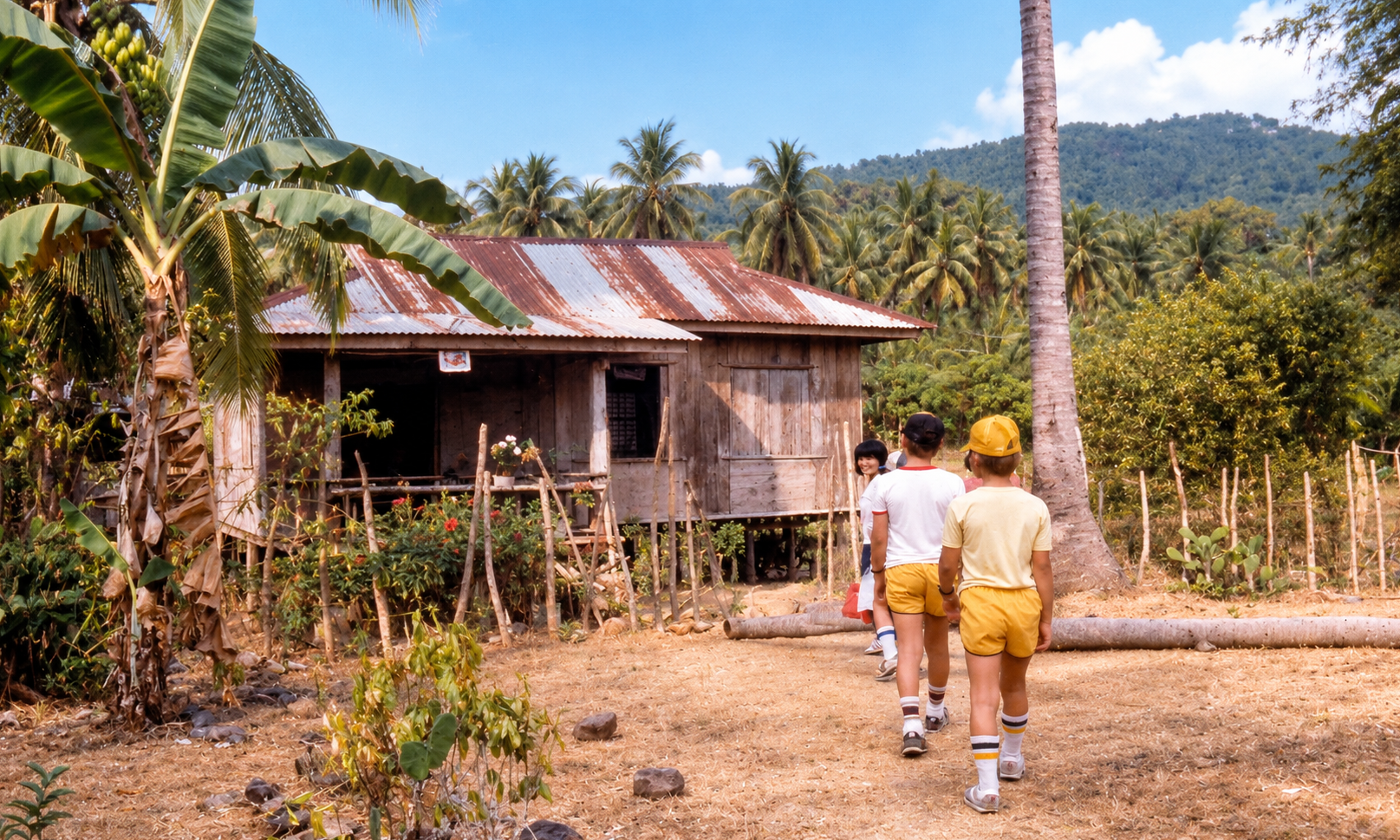 Children walking toward a wooden house surrounded by coconut palms and banana plants.