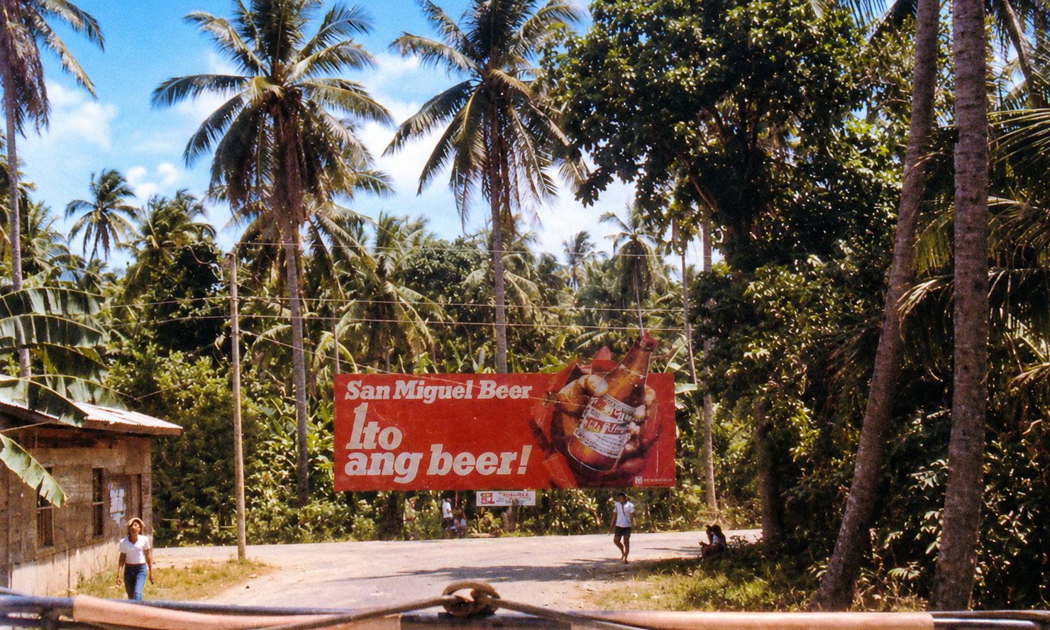 A red San Miguel Beer billboard beside a road surrounded by coconut palms.