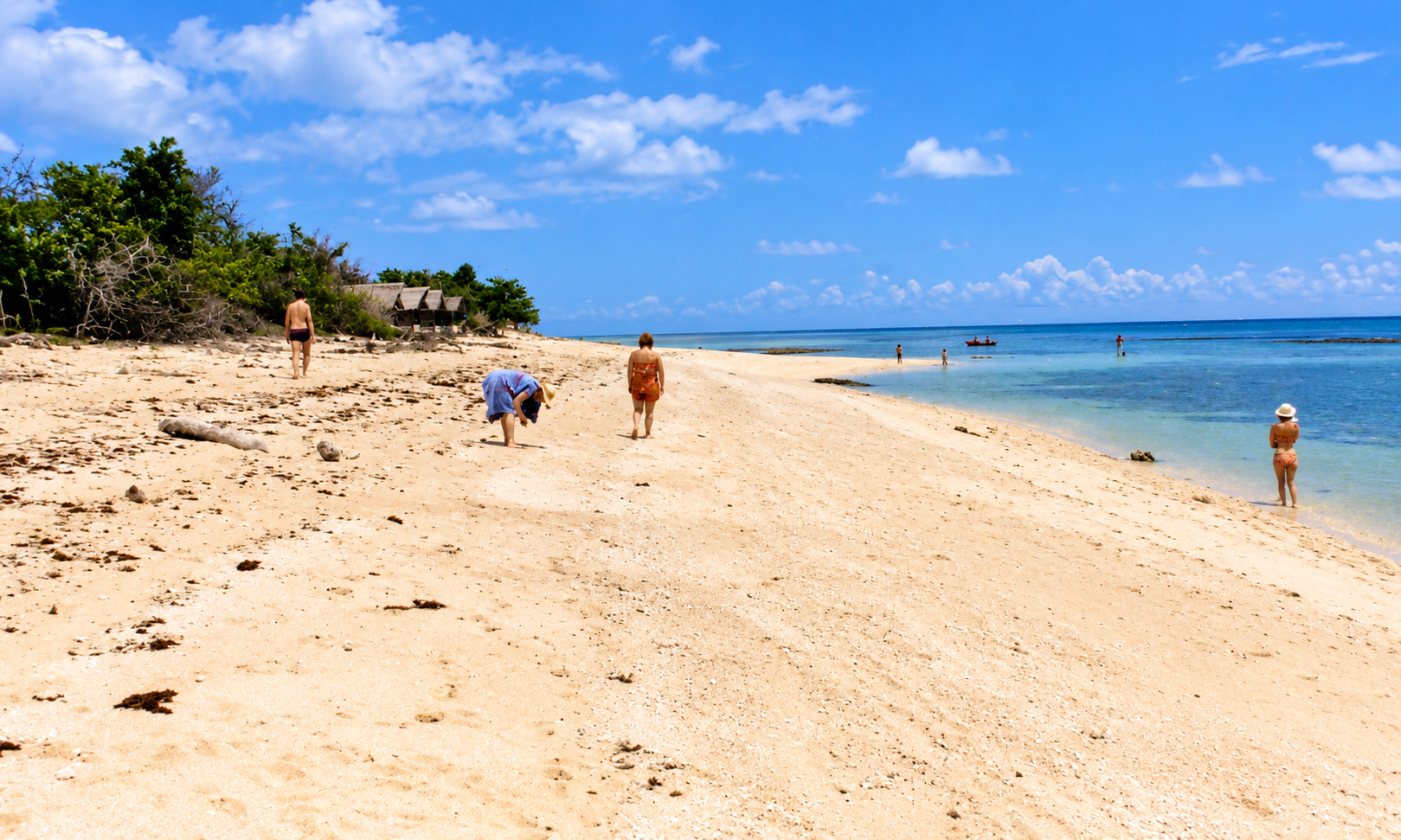 People walking along a wide sandy beach and wading in clear blue water.