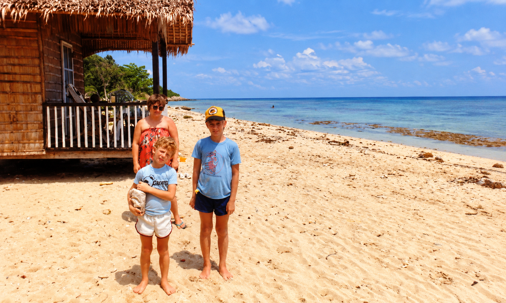 A woman and two boys standing beside a bamboo hut on a white sand beach.