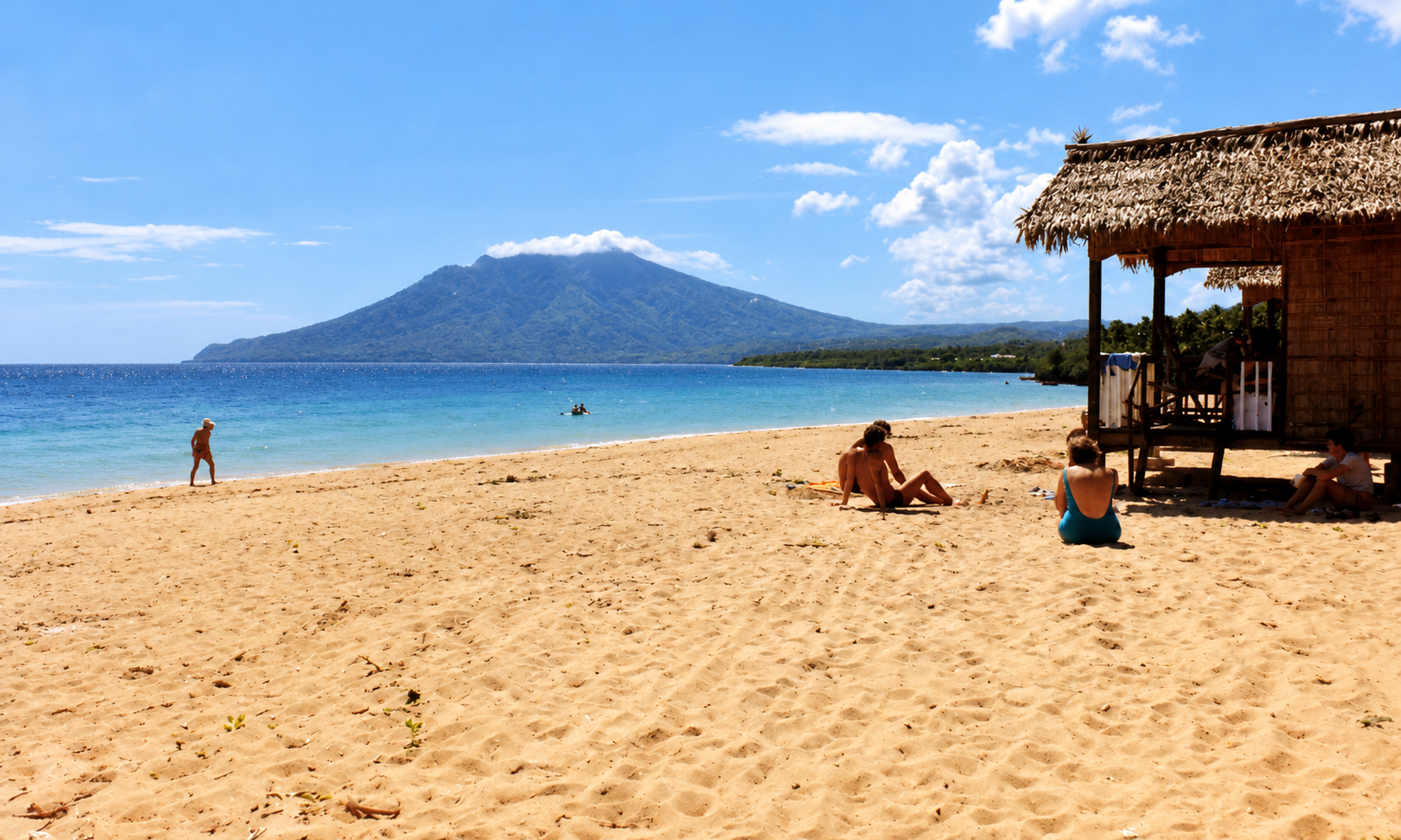People relaxing beside a beach hut overlooking a blue bay and a mountain.