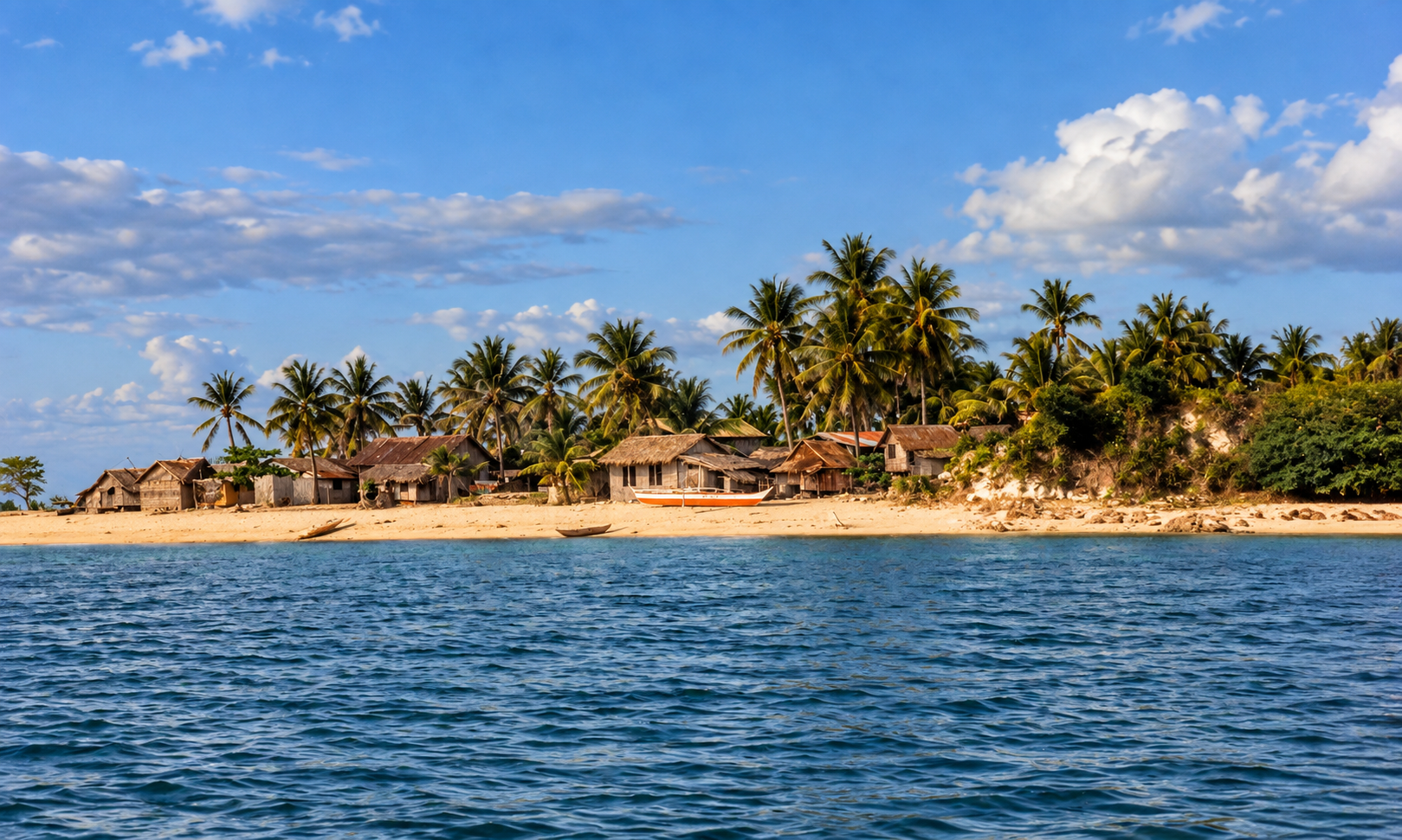 A coastal village of wooden huts and coconut palms seen from the water.