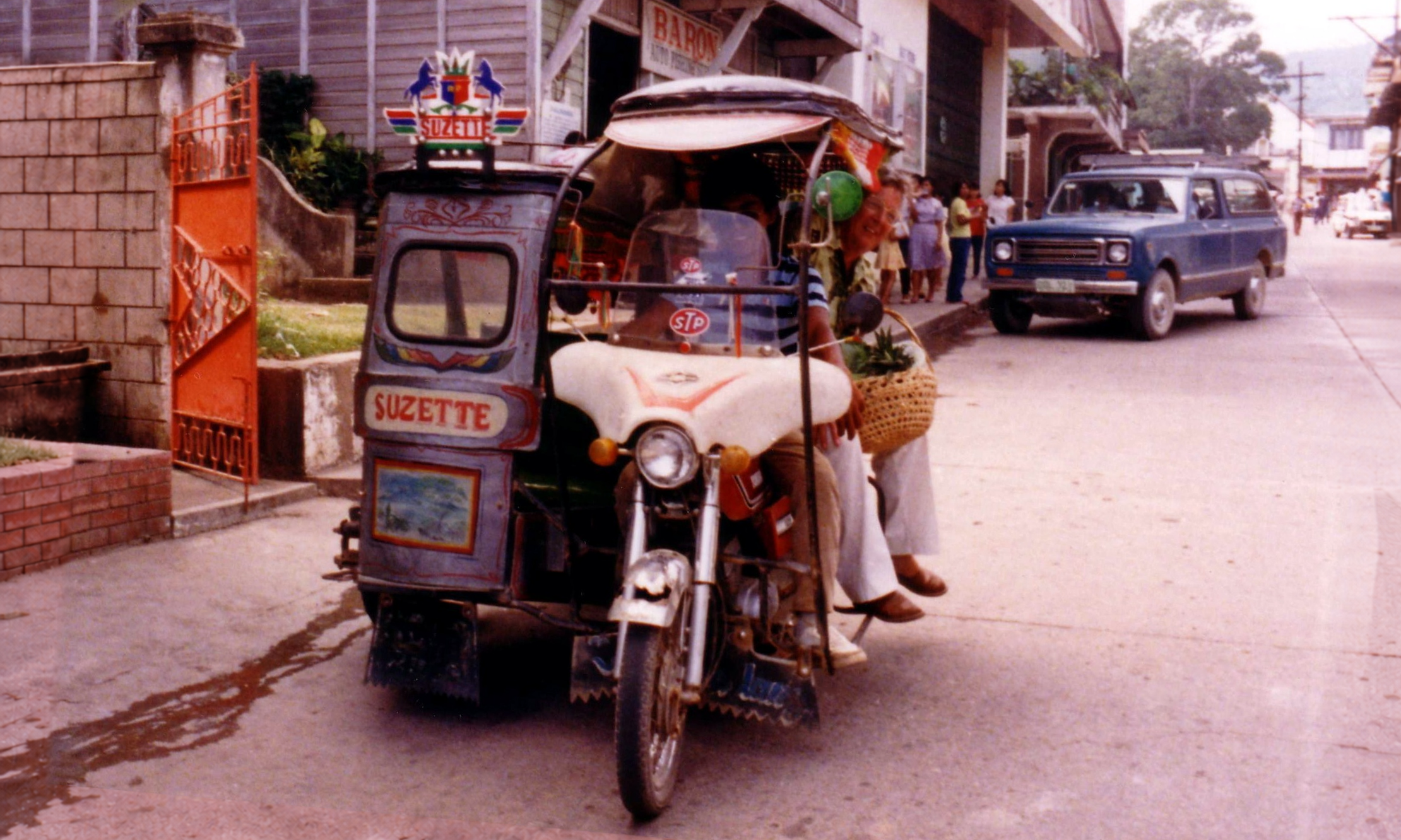 Passengers riding in a motorized tricycle marked Suzette, with a blue car behind them.
