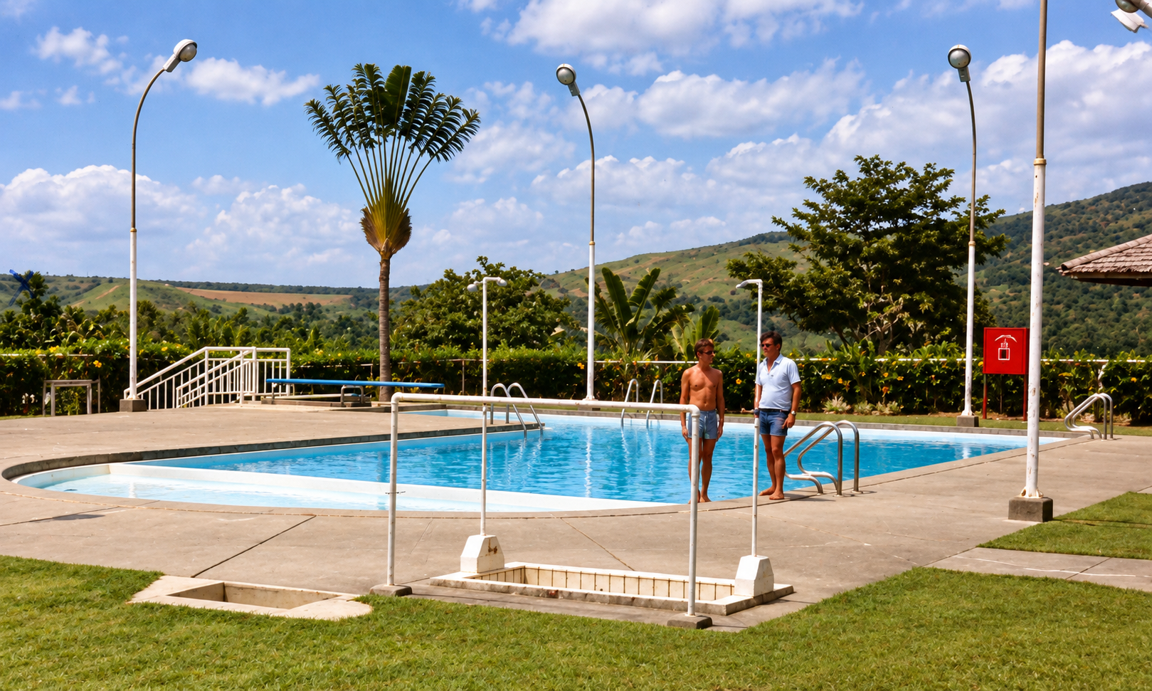 Two men standing beside the blue swimming pool, with tropical trees and green hills behind them.