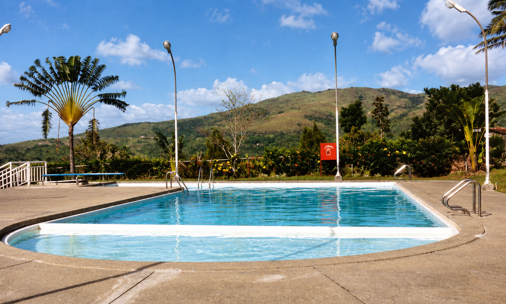 The swimming pool and diving board beneath a blue sky, surrounded by tropical greenery and hills.