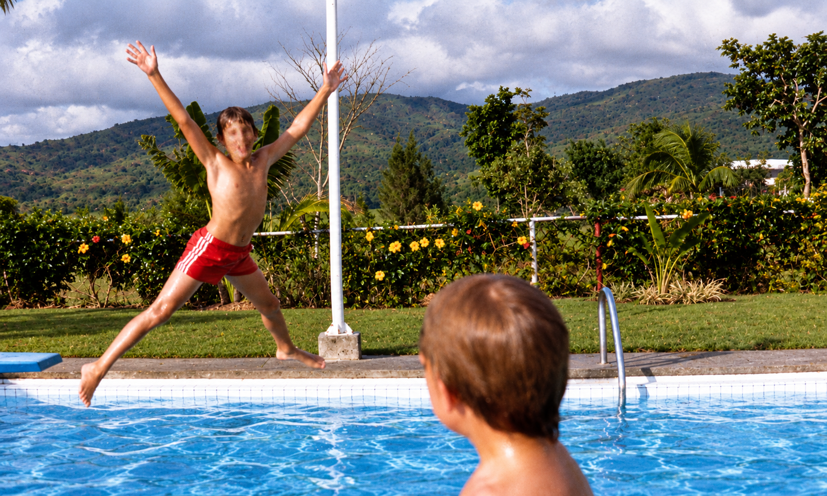 A boy jumping into the swimming pool with his arms spread while another boy watches.