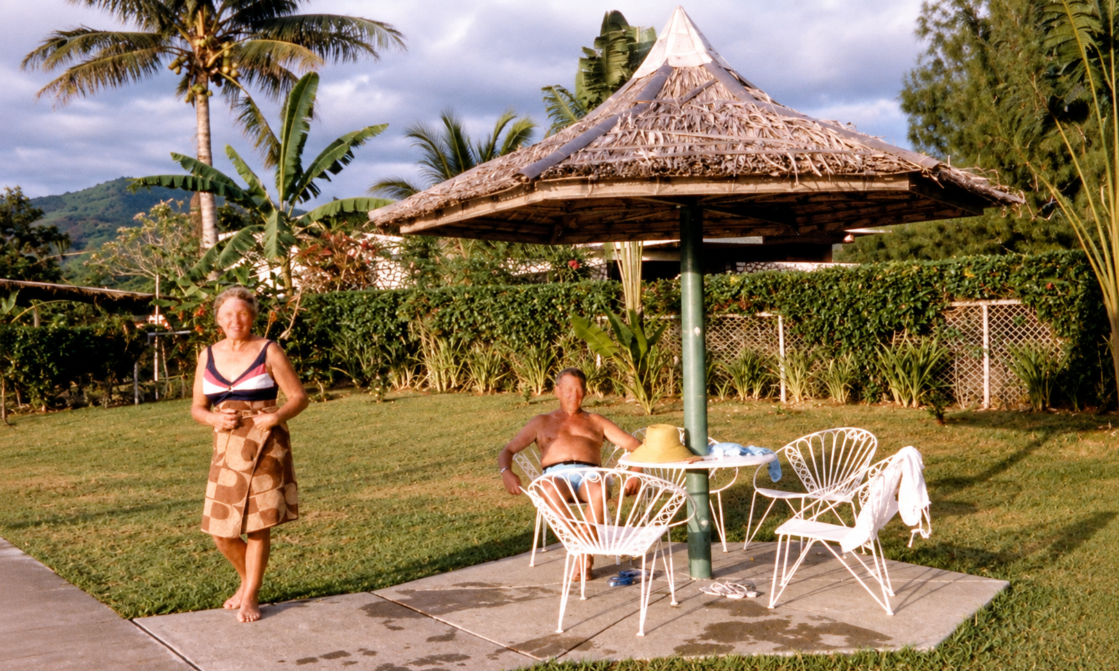 A woman standing beside a man seated under a thatched poolside sunshade.
