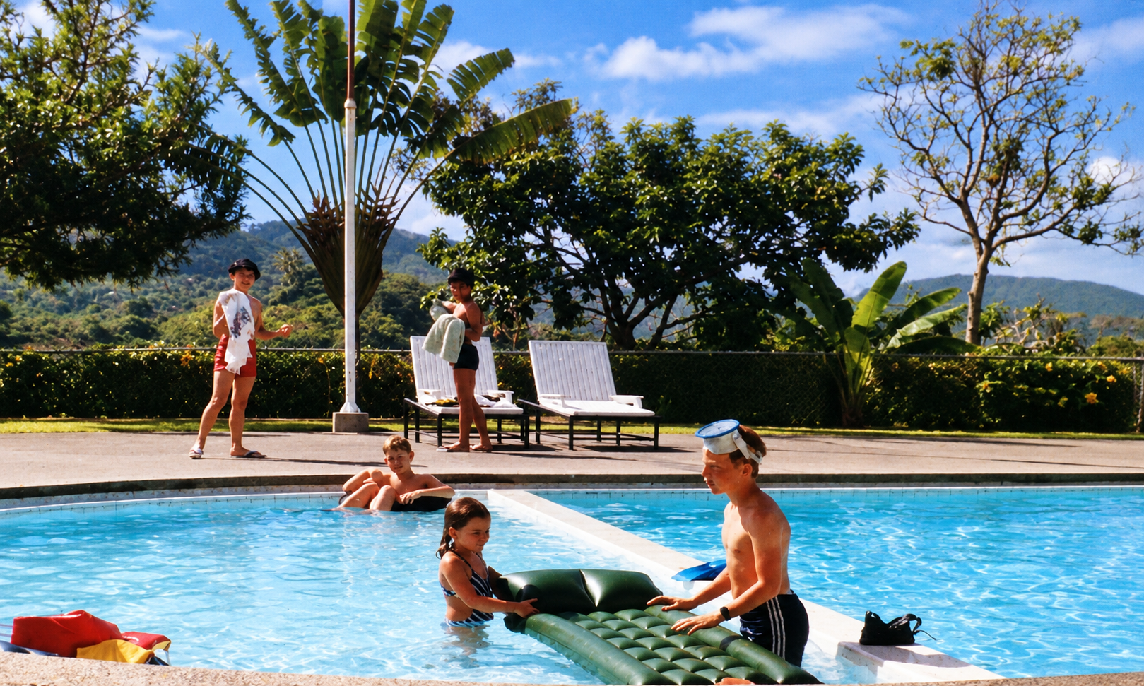 Children playing with an inflatable mattress in the pool while others stand beside the lounge chairs.