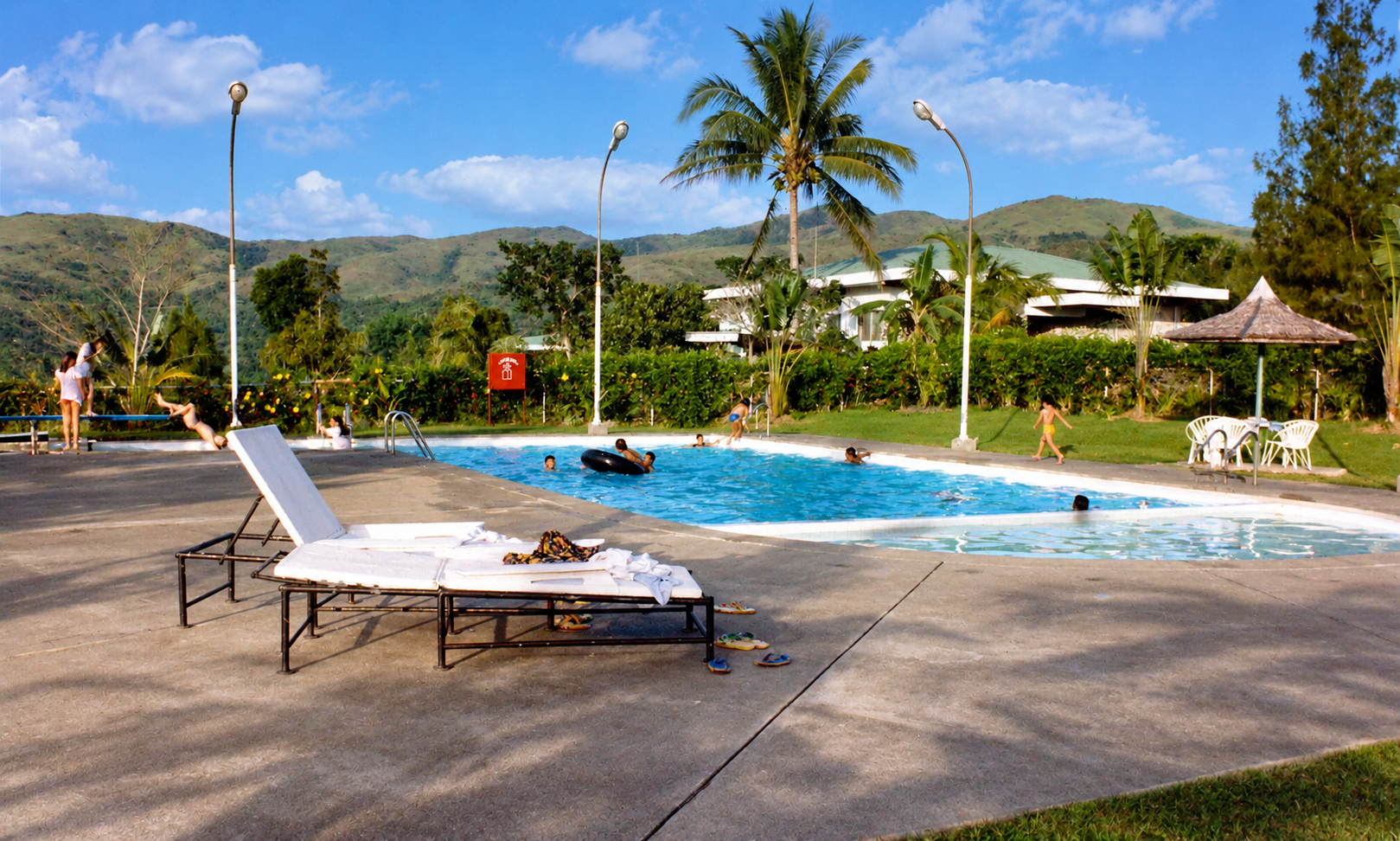 Swimmers and children around the pool, with lounge chairs, a thatched sunshade, and hills behind.